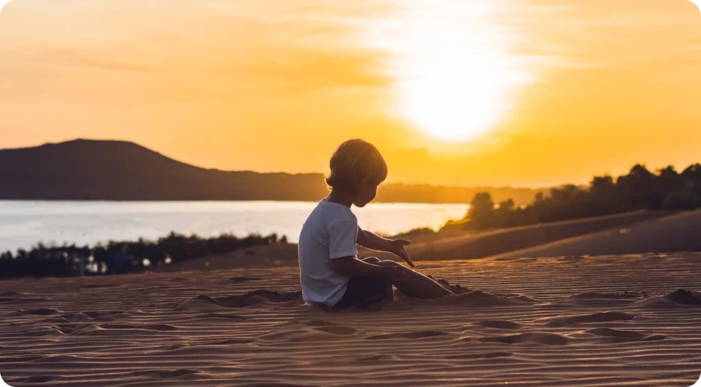 A young child sits on rippled sand dunes at sunset, looking toward a calm body of water and distant hills. The golden sky and low sun create a peaceful silhouette scene with warm evening light.